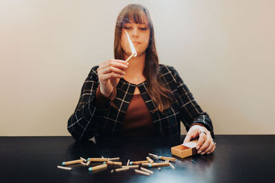 Woman with long brown hair and bangs wears a grid-patterned cardigan while sitting at a black table with several matches strewn about and staring at a lit match she holds.