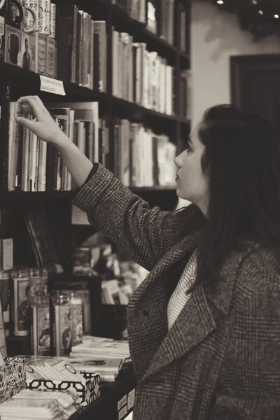 Young woman with very dark brown hair in oversized tweed coat pulling a book off of a bookshelf in the bookstore she is standing in