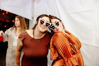 Two women in stylish outfits pose playfully under a tent, captured by Brainerd Lakes photography. They wear heart-shaped sunglasses and smile at the camera, one in a brown dress, the other in an orange pleated top. Soft lighting and another person blurred in the background add warmth.