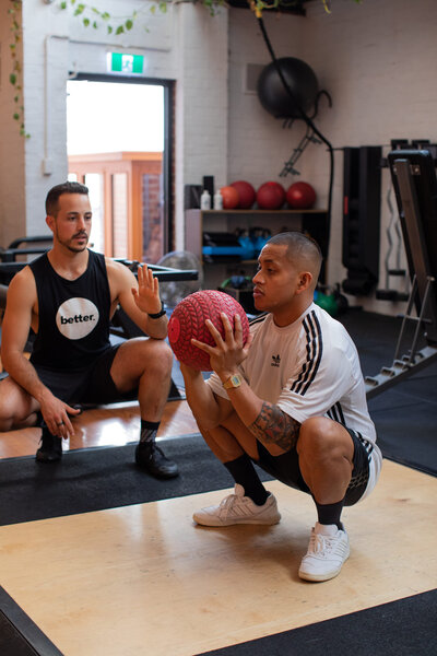 Personal Training with Medicine Ball Squats: A trainer supervising a client performing squats with a red medicine ball in a well-equipped Melbourne gym.