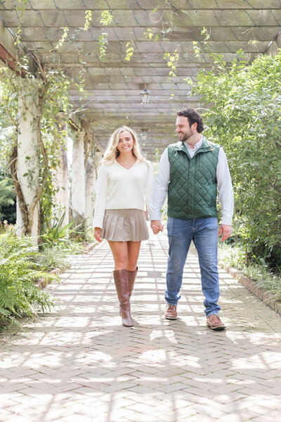 Engaged couple holding hands and walking through a dappled sunlight pergola at Daniel Stowe Botanical Gardens in Charlotte NC