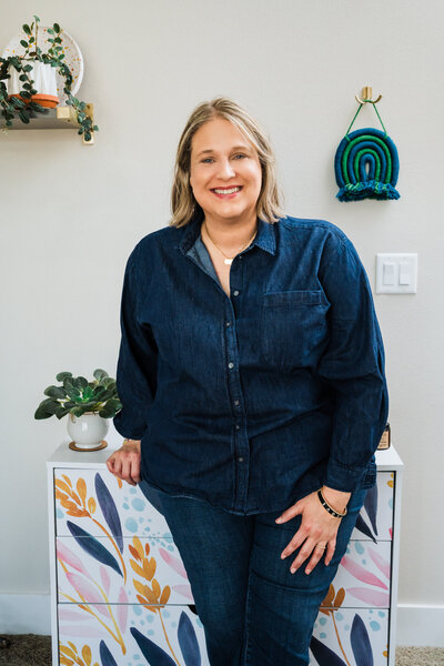 College Coach & Student Success Expert Mya Andersen smiles in a dark denim shirt and jeans, standing by a colorful cabinet with plants and wall art.