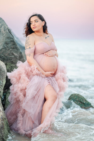 A pregnant woman, wearing a pink tulle dress, stands in the waves at a beach in Virginia. She holds her belly and looks off towards the sunset.