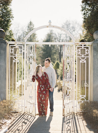 Engagement photograph session at Allerton Park and Retreat Center. In English gardens with tall trees and a white iron gate that the couple is standing in. The gate is large and has double doors. She is wearing a burgundy jumpsuit and looking at him while  holding on to one of the doors. Her other hand is down and holding his hand down by her hip and he is standing behind her looking at her.   Photographed by wedding photographers in Charleston  Amy Mulder Photography