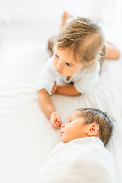 a toddler girl lays on a white bed with her newborn sibling and touches the tip of his nose during their in-home newborn session in Round Rock, TX.