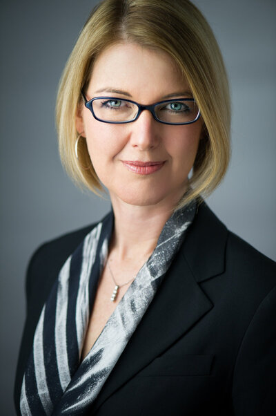 Studio headshot of a caucasian business woman with blonde bob and glasses.