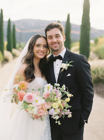 Bride and Groom standing together smiling at the camera for their wine country wedding, she's holding a large pastel colored flower bouquet