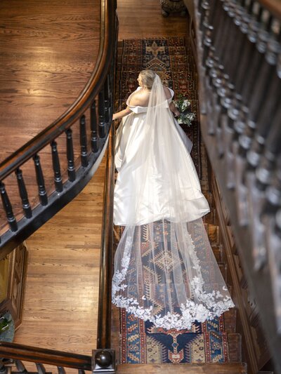 A bride in a white gown with a long lace veil descends a wooden staircase. The scene is viewed from above, and the staircase has ornate railings. She holds a bouquet, and a patterned rug is visible on the steps.