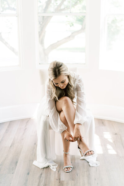 A woman in a white dress sits on a chair near a window, bending down and adjusting her shoe strap, meticulously preparing for her special day with the help of a renowned Canadian wedding planner.