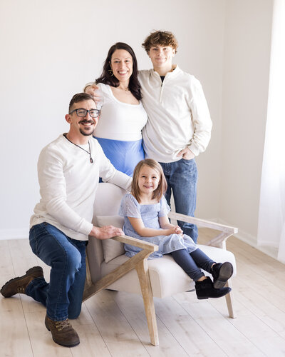 Family of four in a studio, smiling at the camera. Dad is knelt down next to their young daughter who's sitting in the chair.  Mom and son are standing up next to each other behind the chair.