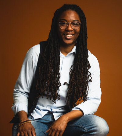 Black woman with long locs and glasses, wearing a light colored button up shirt smiling at the camera in front of a reddish orange backdrop
