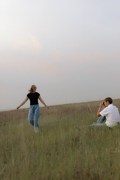 A man takes a photo of his girlfriend as she twirls in a grass field.