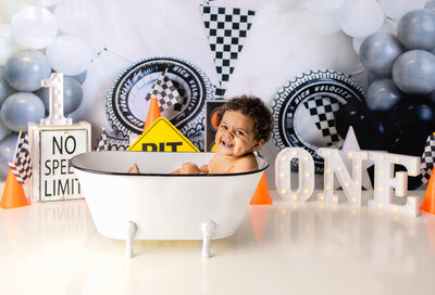 Smiling baby boy in a mini bathtub during race car themed cake smash bath session with checkered flags in Waldwick NJ studio