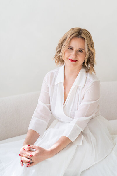 žena v bílých šatech sedí na světlé pohovce a usmívá se