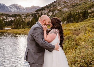 Bride and groom smiling and holding each other close by a lake with golden foliage and alpine peaks during a fall elopement in Breckenridge, Colorado