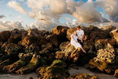 Woman looking at man sitting on the rocks of a jetty at the beach during a Tampa engagement photography session.