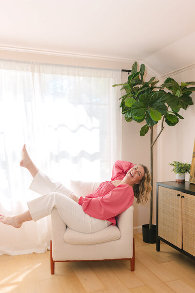 Mollie Mason in a pink top and white pants laying sideways in a white chair with her legs up and smiling