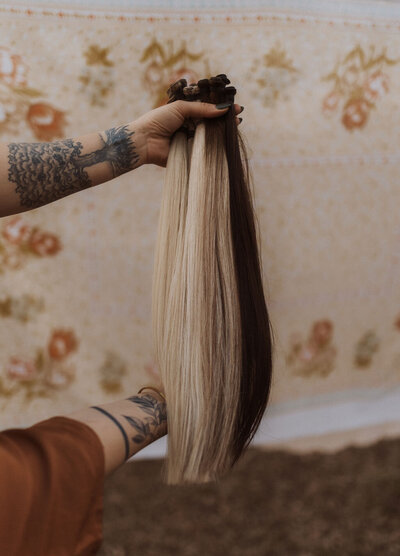 The best hair extensions professional in Frisco is holding up her flagship product. The professional has palm trees, leaves and leaves tattooed on her arms. Behind the hair extensions is a wall tapestry with flowers