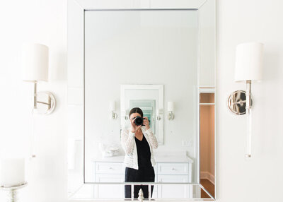 A woman lifestyle photographer taking a picture of herself in the mirror with a camera.