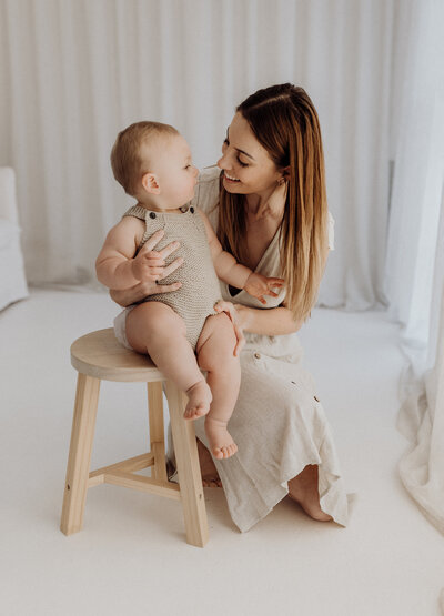 A mother holds her little baby while she sits on a stool and looks at one another while lucy jane photographs them in her christchurch photography studio