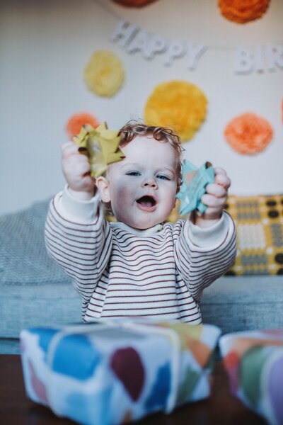 A toddler in a striped jumper holding up two bows from a birthday present in front of him