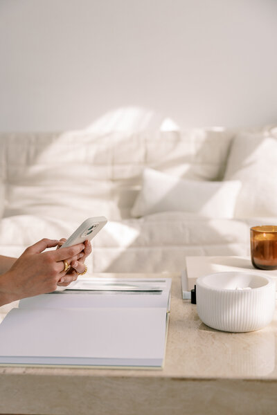 Ein Sofa im Hintergund. Vorne sitzt eine Frau und sie hat ein Handy in der Hand. Vor ihr liegt ein Notizbuch auf dem Tisch.