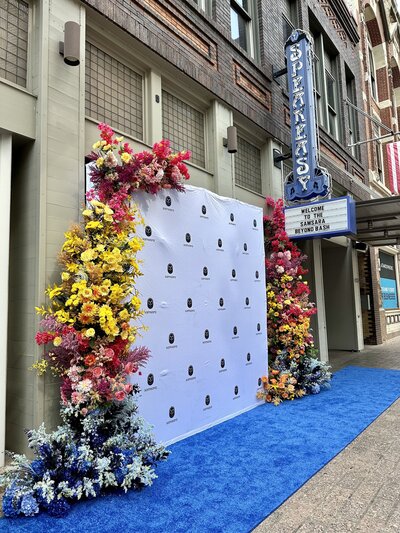 Corporate event photo backdrop with bright floral installation and blue carpet, styled for high-impact brand moments in Austin.
