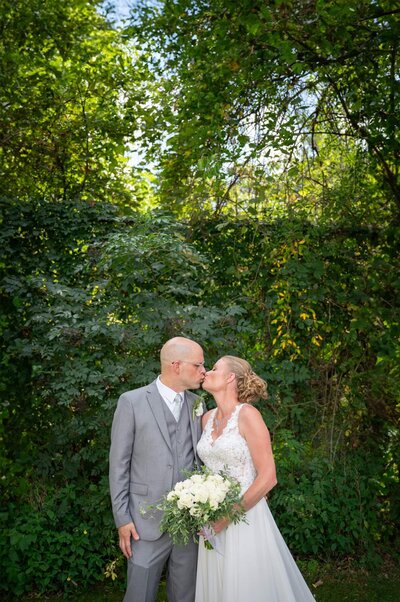 bride and groom kiss in front of ivy covered fence and trees