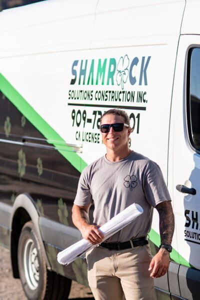 Man in a grey shirt standing in front of a Shamrock Solutions Construction Van. He is holding  construction plans and smiling.