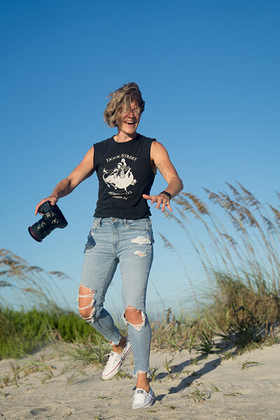 A photographer holds her camera in one hand and laughs while her hair covers her face with beach dunes and tall grasses in the background. 