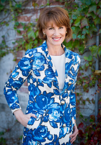 Headshot photo of a beautiful travel agent wearing a blue and white floral jacket and smiling in front of a brick wall with greenery in San Juan Capistrano, CA.