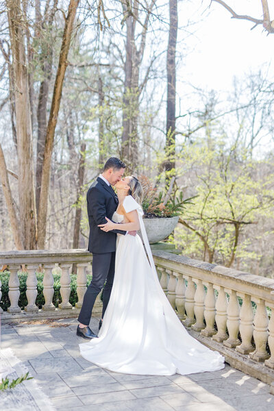 A bride and groom kiss as they stand outside with blooming spring trees around them. He is wearing a black tux.