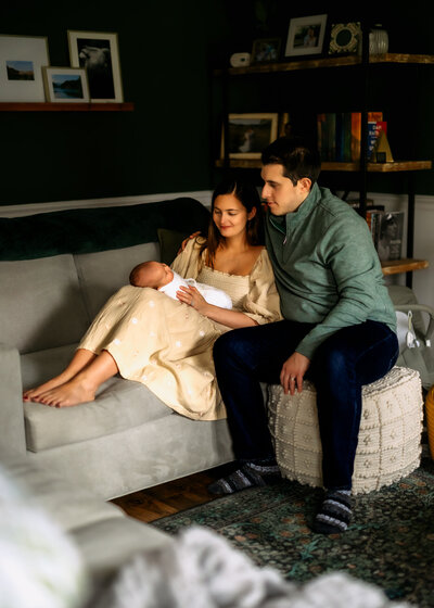 Mom is sitting with her legs up on a tan couch with baby laying with their back against her thighs and dad is sitting on an ottoman with his arm around her in a loving embrace. Baby is asleep with a white blanket around them while mom and dad look down at them smiling. They're in a living room with bookcases behind them for this newborn photography session.
