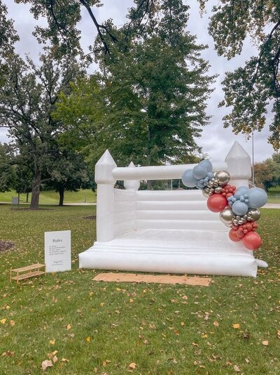 A white bounce house set up outside with a blue, gold, and red balloon garland attached to the front of it.