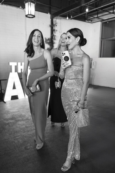 Three women in elegant evening dresses walk together, smiling and holding clutches. The woman on the right is holding a smartphone with a prominent accessory attached. They are indoors, passing by illuminated letters in the background.