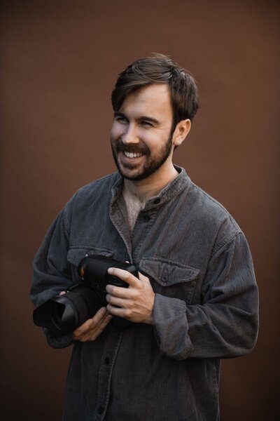 Photograph of Tom Keenan the photographer holding his Nikon D850 in front of a brown backdrop in Cambridge