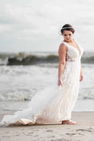 A radiant bride walks barefoot along the shoreline, her lace wedding gown flowing effortlessly in the ocean breeze. With a serene expression, she embraces the moment, fully present and stress-free on her wedding day. This dreamy beachside portrait captures the essence of Alabama wedding photography—focusing on timeless elegance, natural beauty, and the joy of a relaxed, unforgettable celebration.