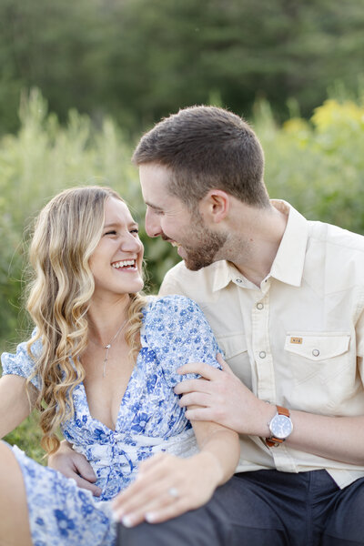 Engaged couple smile at each other while they sit in a field