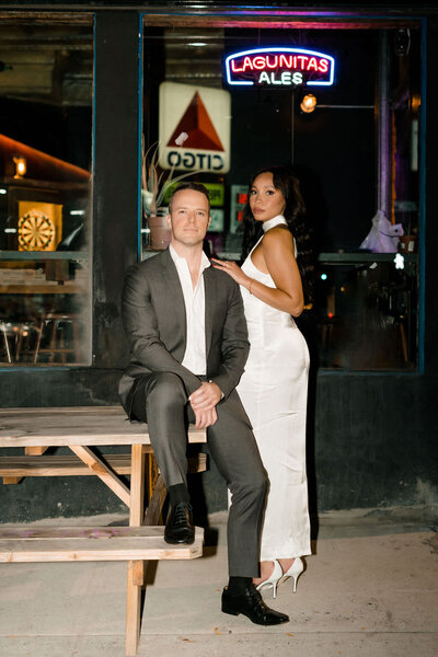 groom sitting on a picnic table outside at night with his bride standing next to him resting her arm on his shoulder