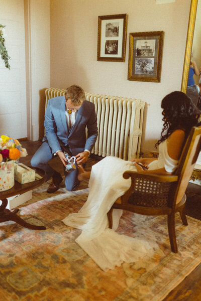 Groom helping bride put on her wedding shoes before the ceremony, captured in an intimate documentary style by Emily Ambie.