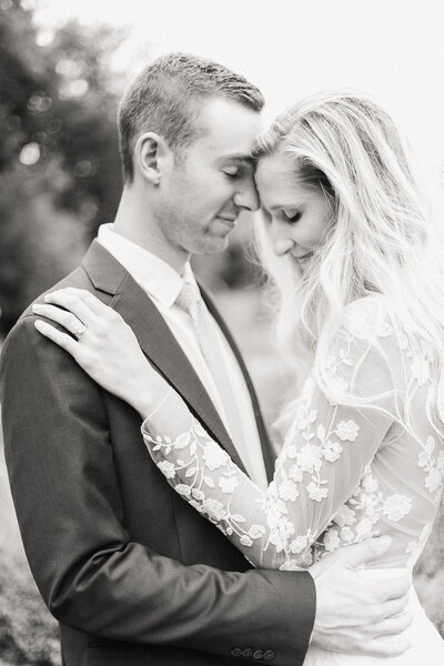 A black and white portrait of a bride and groom embracing, highlighting the bride's long-sleeved lace gown.