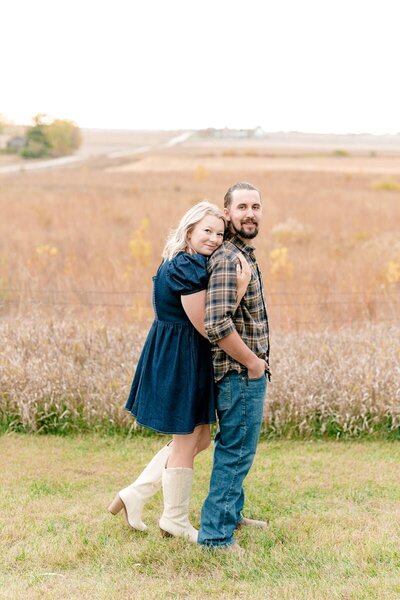 A couple sharing a loving embrace under a beautiful archway, captured during their engagement photo session. The woman is wearing a light blue dress, and the man is in a casual shirt and pants, kissing her cheek affectionately.