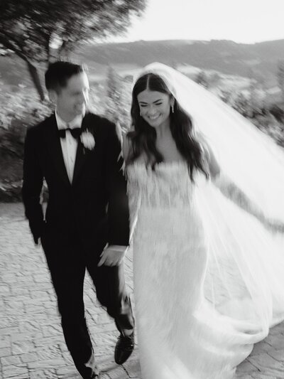 A black and white image of a couple in wedding dress runs through the vineyards