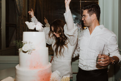 Bride dancing with arms in air next to groom in front of white and coral wedding cake