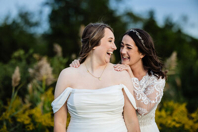 Two brides embracing tenderly in their white gowns against a backdrop of a scenic sunset and rural landscape at Allen Hill Farm
