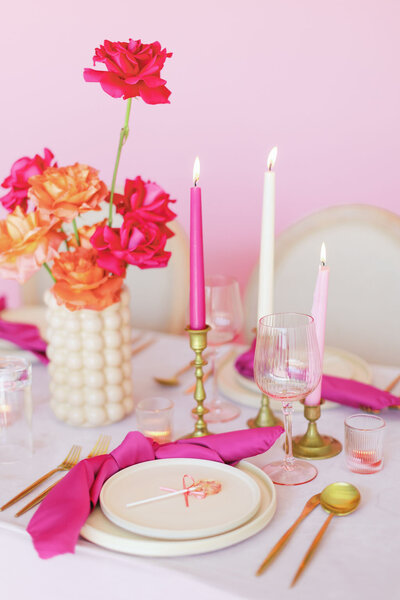 Bright pink themed table setting with white plates, gold silverware, pink napkins and candles, and pink and orange flowers in a white vase