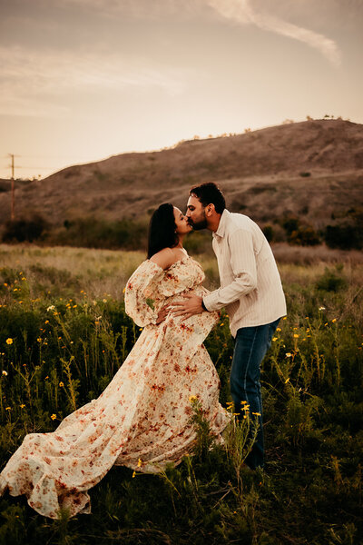 Expecting couple kissing in a wildflower field, with the woman wearing a flowing floral gown during a romantic sunset maternity session in Southern California, photographed by Brittani Luce

