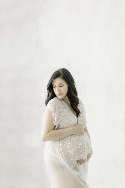 A maternity photo taken in a Dallas photography studio of an expecting mother holding her belly while she’s dressed in an elegant lace robe.