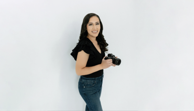 Lissette Houston Photographer standing infront of a light grey background holding a camera.