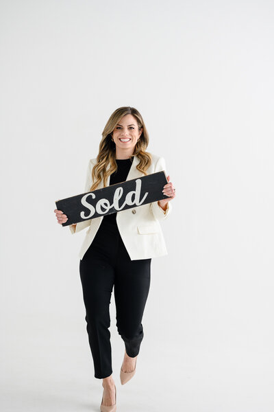 A brand photographer in Denver takes a photo of a real estate agent in a white suit walking toward the camera with a sold sign in her hands for her branding photo shoot for brand photography.